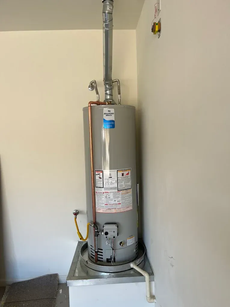 A gas water heater standing on a metal drip pan in a corner with copper pipes, a vent, and a gas line connection.