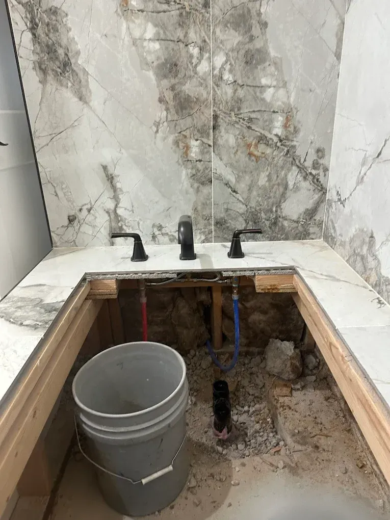 A bathroom vanity under construction with marble countertop, black faucets, and exposed plumbing over an open cabinet.