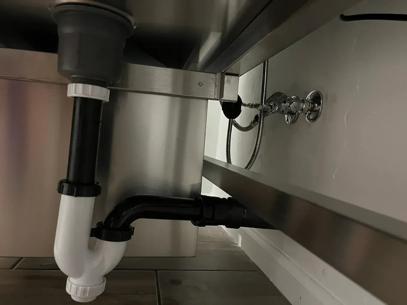 Under-sink plumbing showing a white P-trap connected to black piping and stainless steel sink basin.