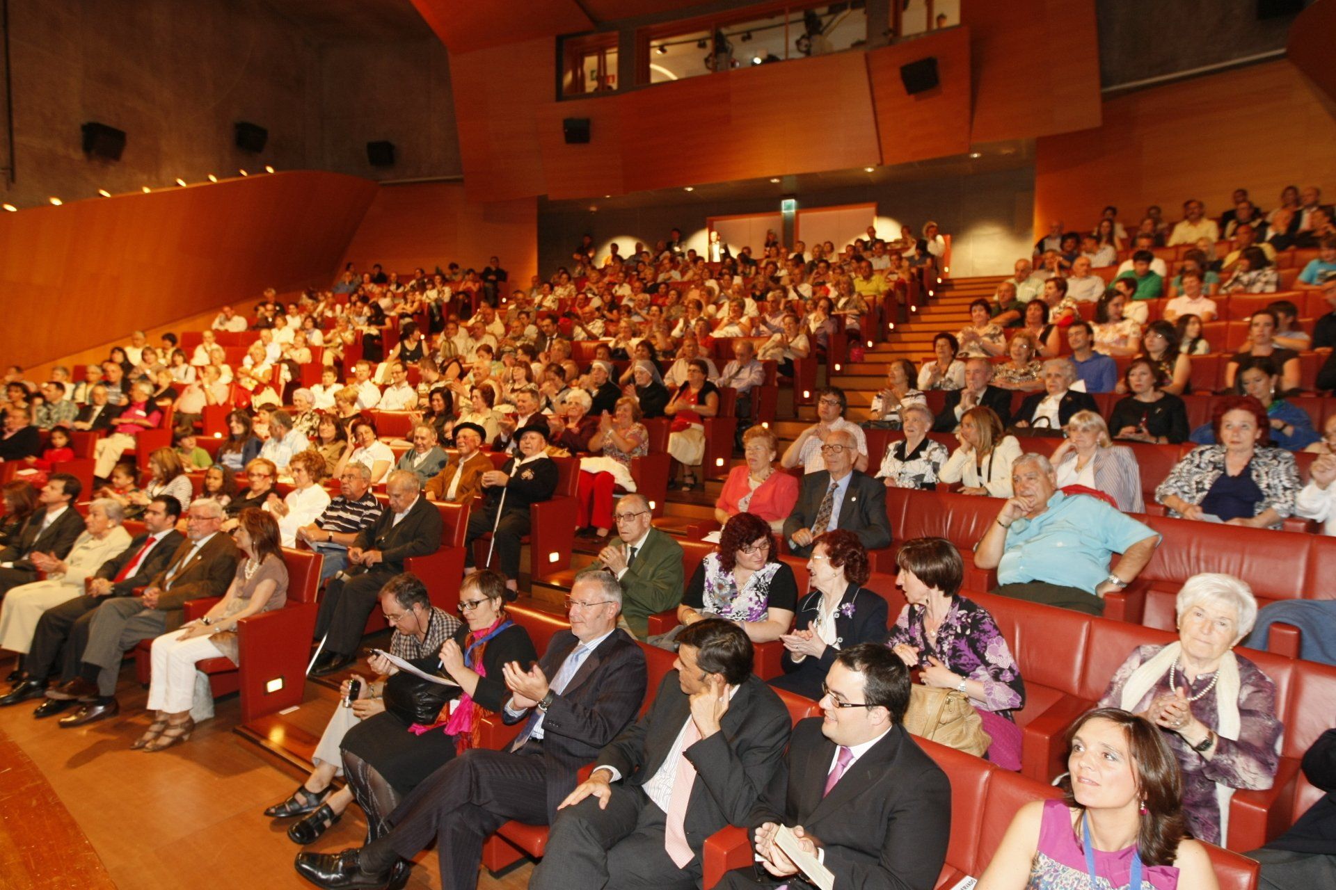 Público en butacas rojas de teatro, asistiendo a un evento. Se ven personas de diversas edades y vestimentas.