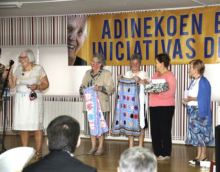 Grupo de mujeres mayores sonriendo, con regalos, en una ceremonia. Detrás de ellas hay una pancarta amarilla con texto.