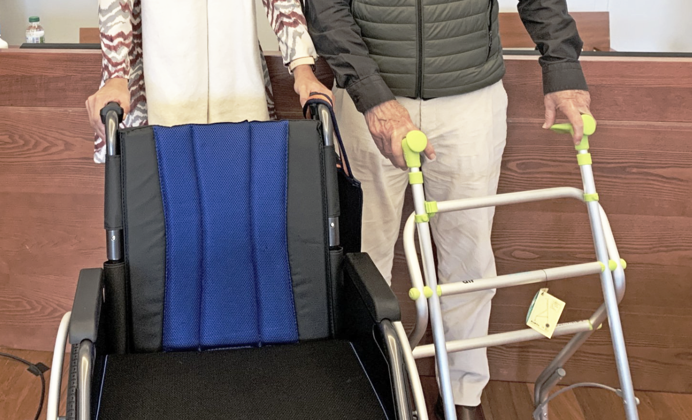 Una persona con un andador y una silla de ruedas. Paneles de madera al fondo.