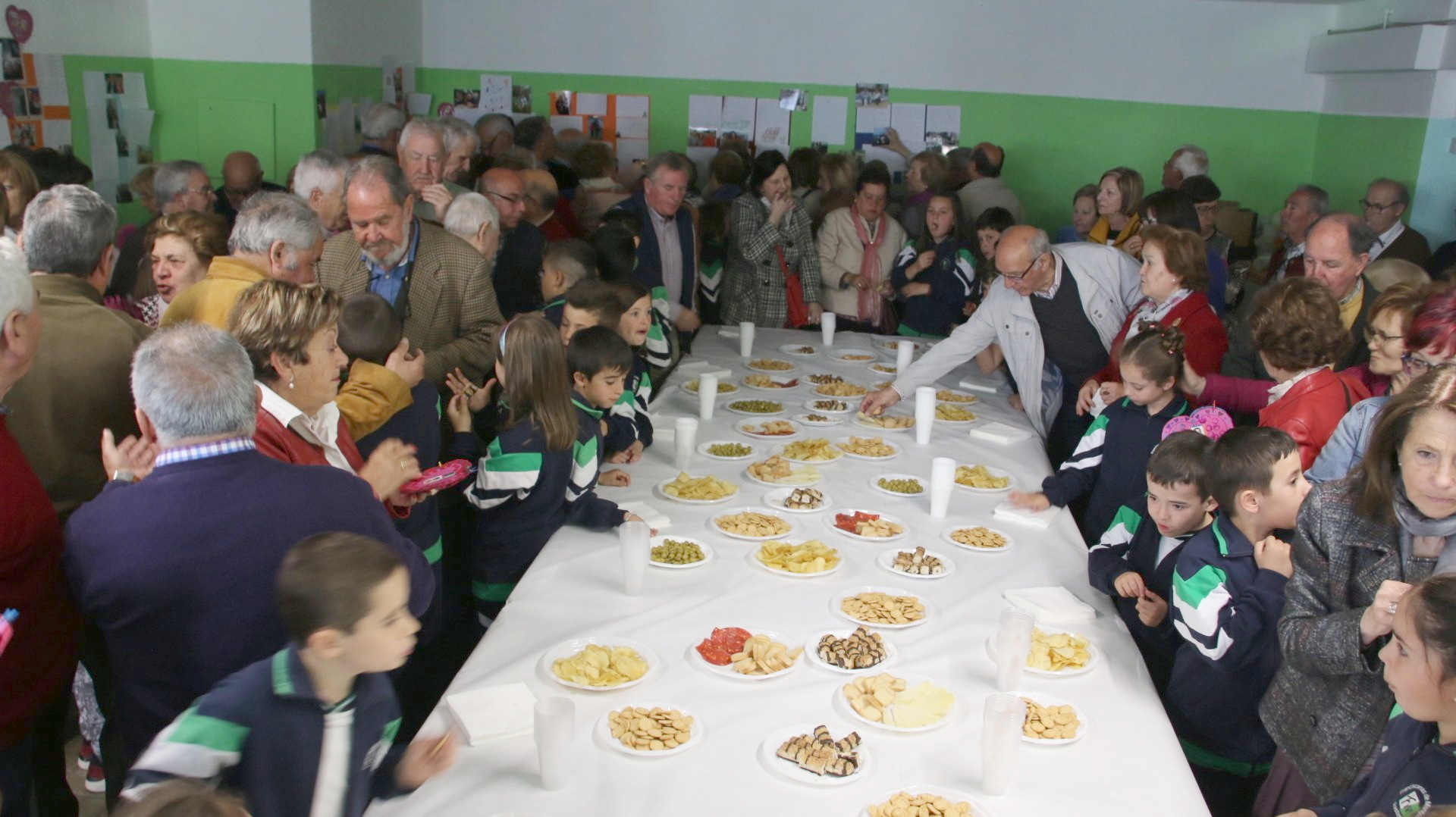 La gente y los niños se reúnen alrededor de una mesa cargada de comida en una habitación bien iluminada.