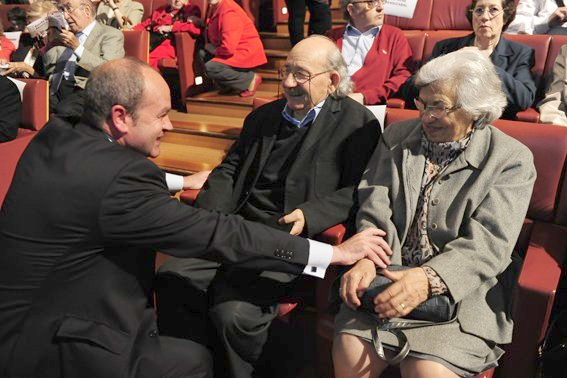 Un hombre de traje saluda a una pareja de ancianos en un auditorio. El hombre toca el brazo del hombre sentado y el hombro de la mujer.