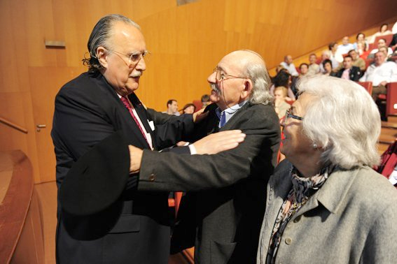Tres personas se abrazan en un auditorio. Dos hombres y una mujer con atuendo formal. Hay gente sentada al fondo.