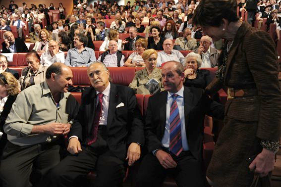 Hombres de traje conversan en un auditorio; el público está sentado detrás.