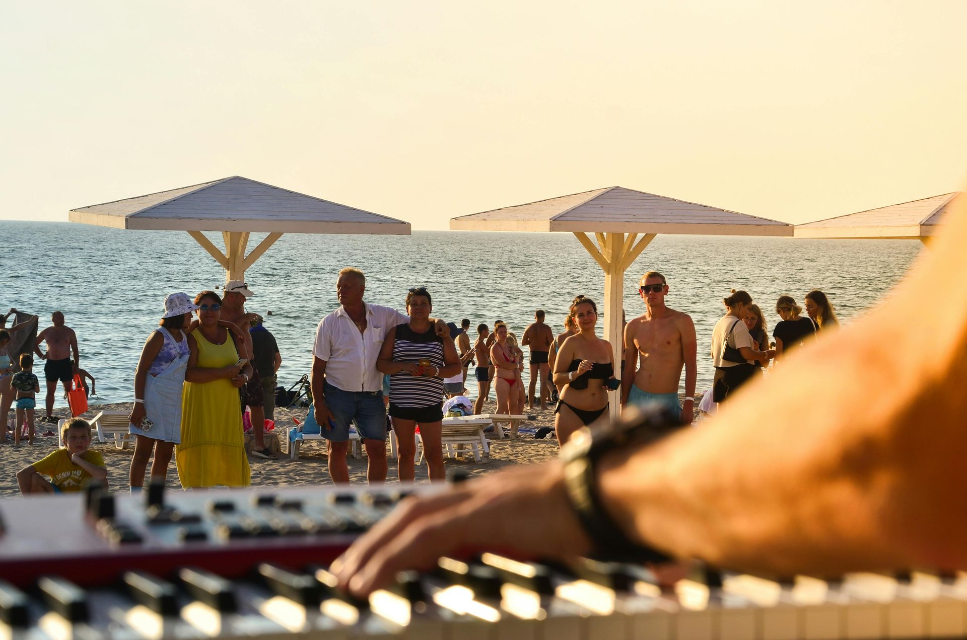 A man is playing a keyboard on the beach while people are standing under umbrellas.