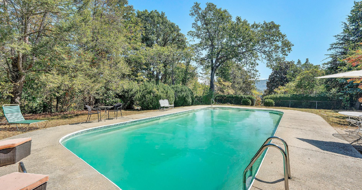 Outdoor swimming pool with turquoise water, surrounded by trees and a stone deck under a clear sky