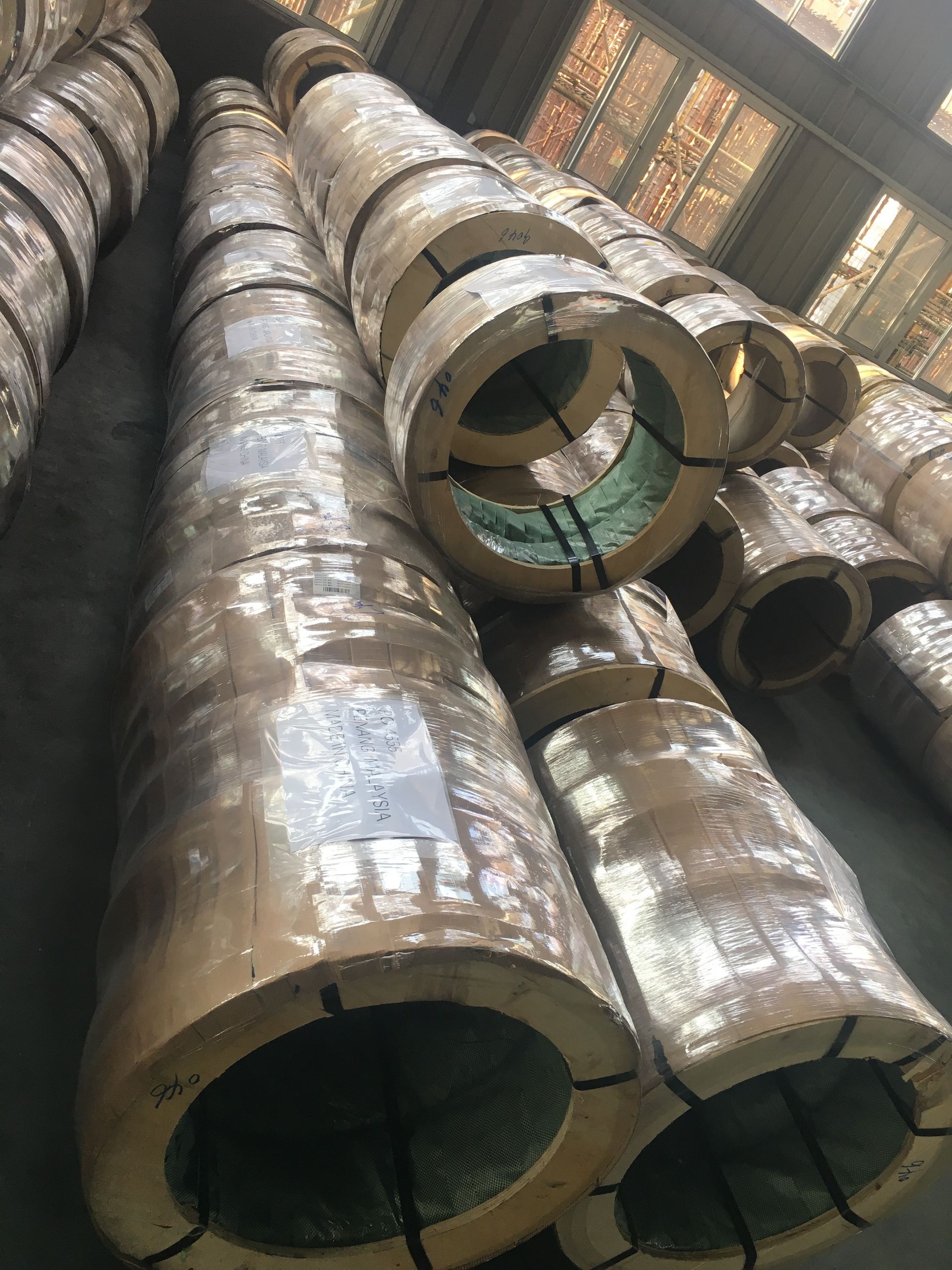 Coils of metal, wrapped in protective plastic, stacked in a warehouse.