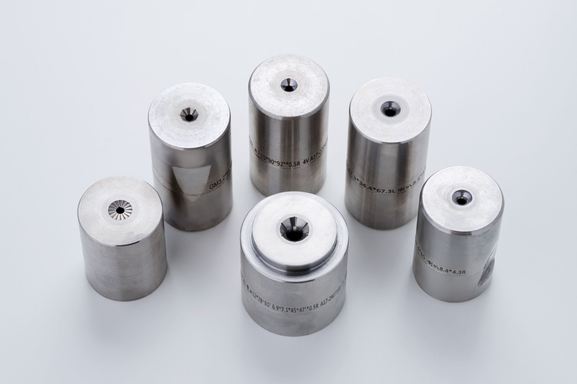 Six cylindrical metal dies arranged on a white surface, each with a central hole and etched markings.