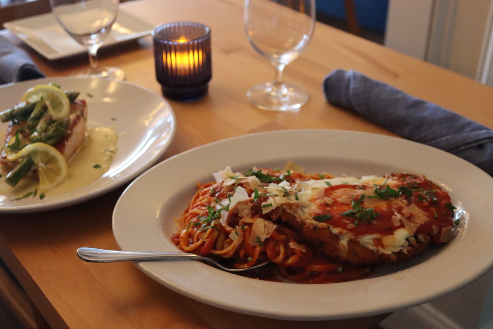 a plate of pan seared salmon and chicken parmesan are sitting on a wooden table with a candle and glasses