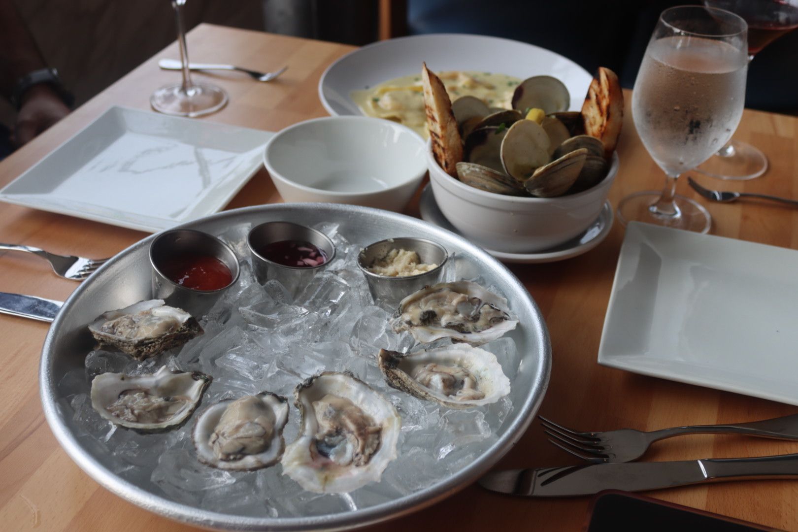 A tray of oysters on ice, clams in garlic sauce in a bowl and lobster ravioli on a white plate sitting on a wooden table
