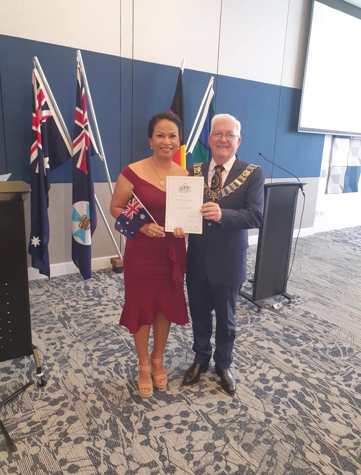 Woman in Red Dress Holding Certificate, Man in Suit, Flags, Room — Immigration Agents in Cairns North, QLD