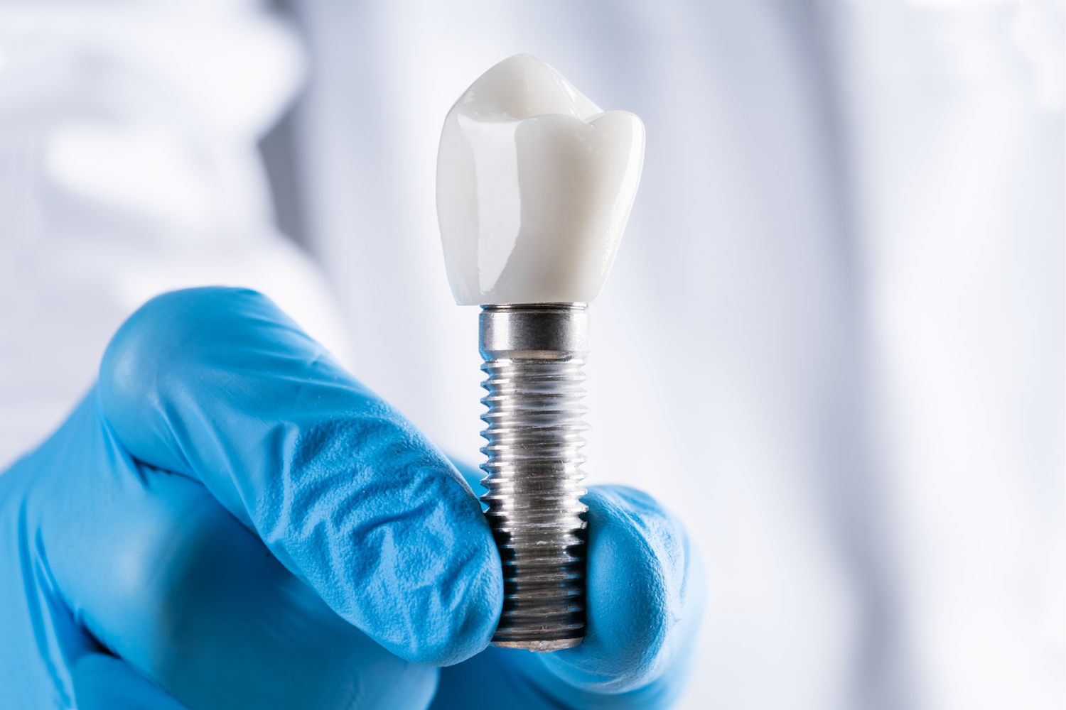 Dental implant held in gloved hand. The implant has a metal screw base and a white ceramic tooth crown.