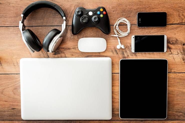 Headphones, game controller, laptop, tablet, mouse, earbuds, and smartphones on a wooden surface.
