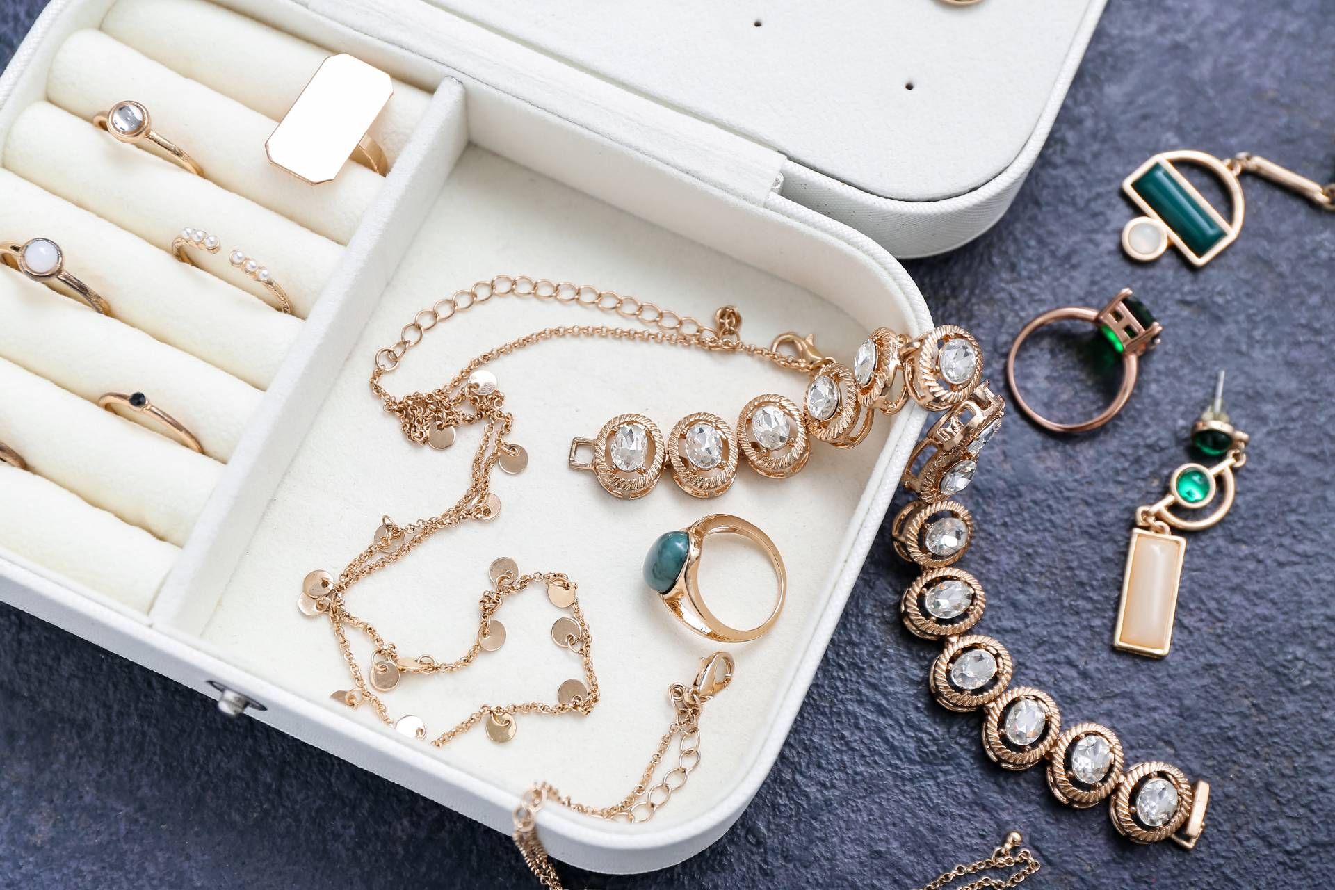 White jewelry box with various gold necklaces, rings, and earrings on a dark grey surface.