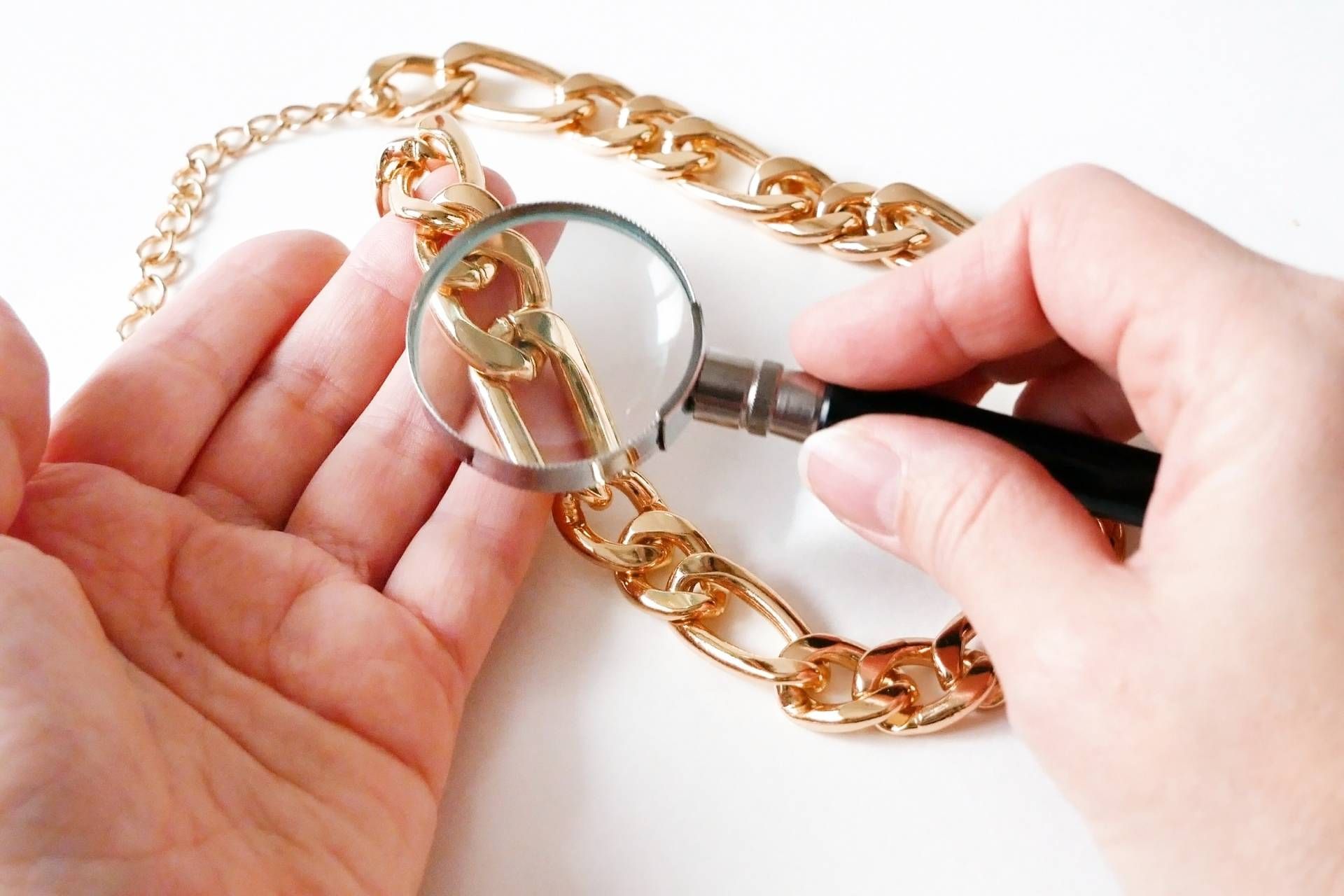Hands holding gold chain, examining it closely with a magnifying glass on a white surface.