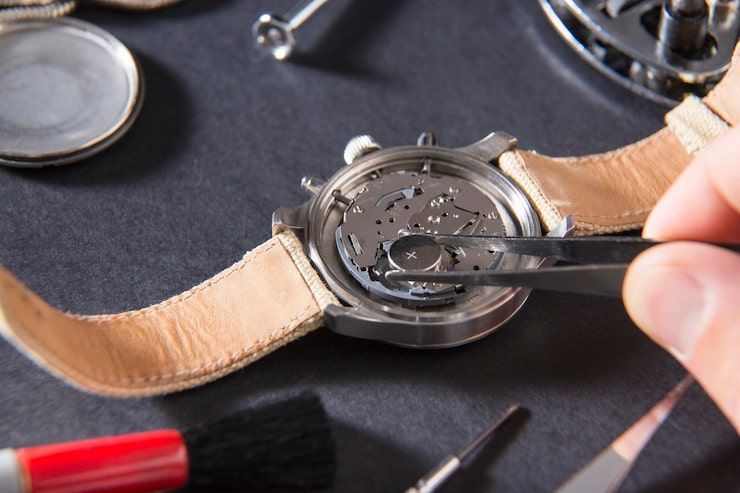 Watch being repaired with tweezers; tools and parts on a dark surface.