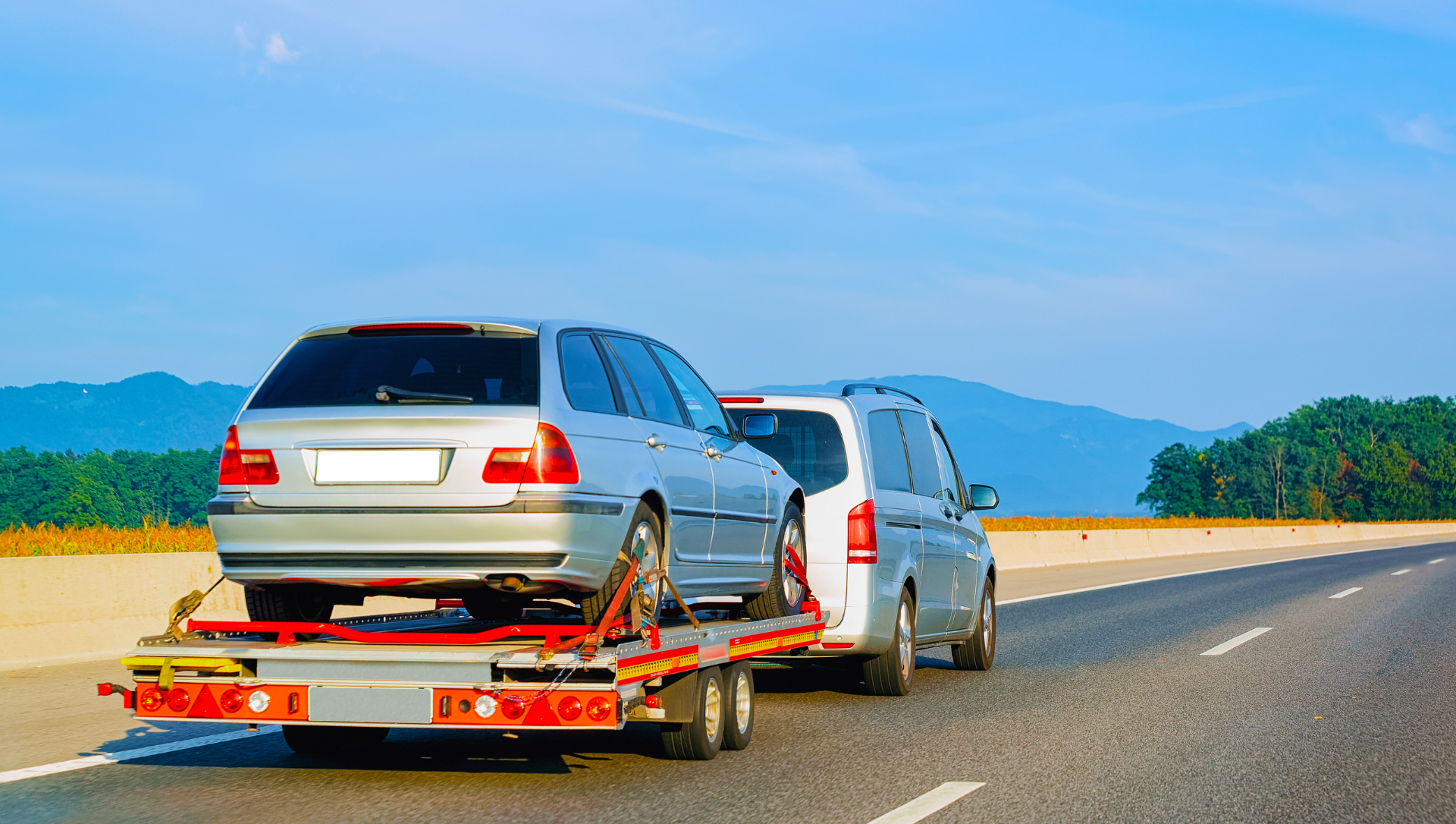Car being towed on a flatbed trailer on a highway. Blue sky, green hills in the background.
