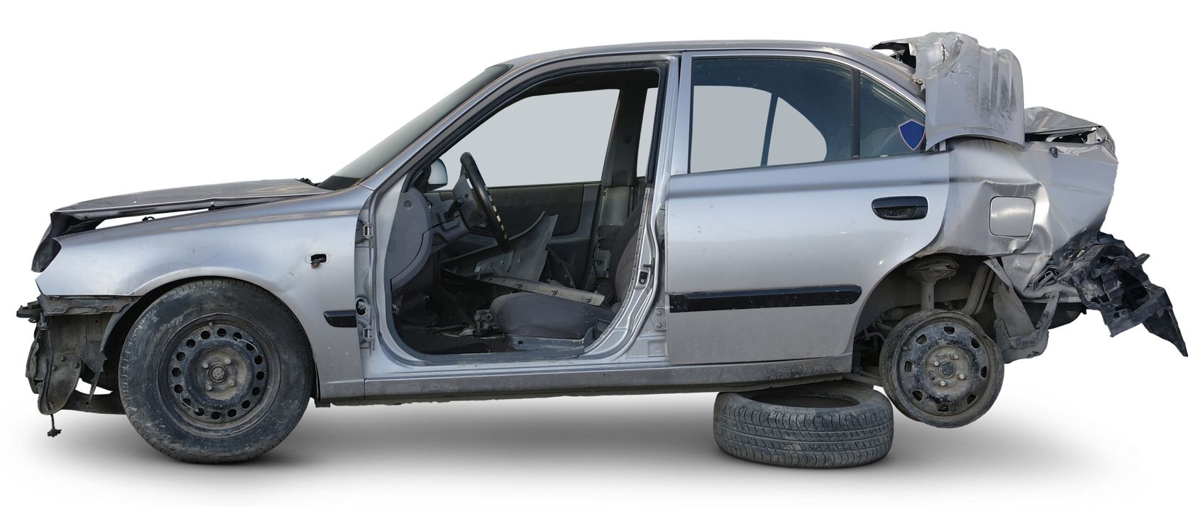 A severely damaged, silver sedan, missing parts, sitting on a tire, likely a junked car.