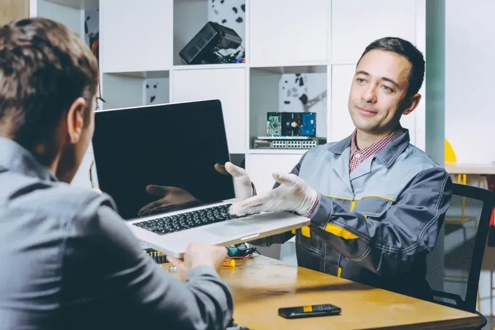 Man in Grey Uniform Handing a Laptop to Another Man at a Desk. Office Setting — Micromac Computers in Charmhaven, NSW
