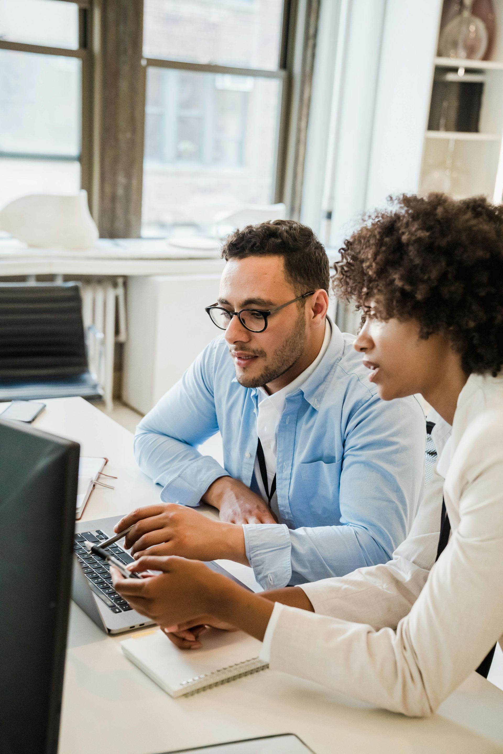 Man and woman looking at laptop screen, reviewing documents together. Bright office setting.