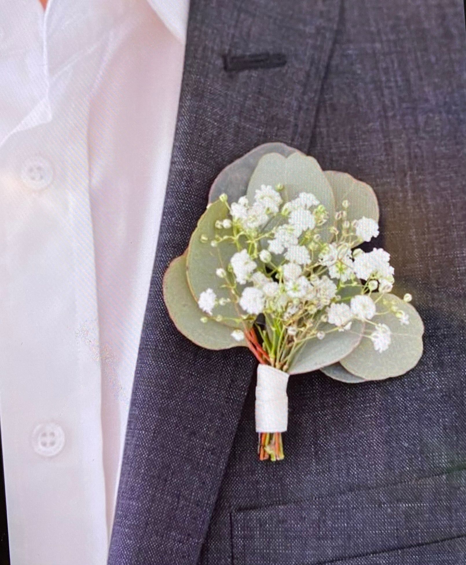 A groom is wearing a buttonhole with baby 's breath and eucalyptus leaves.