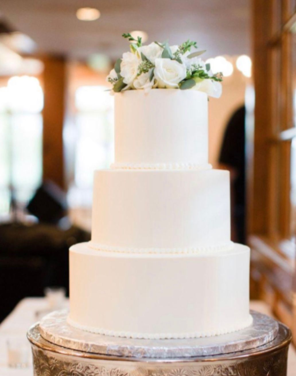 A white wedding cake is sitting on top of a silver cake stand.