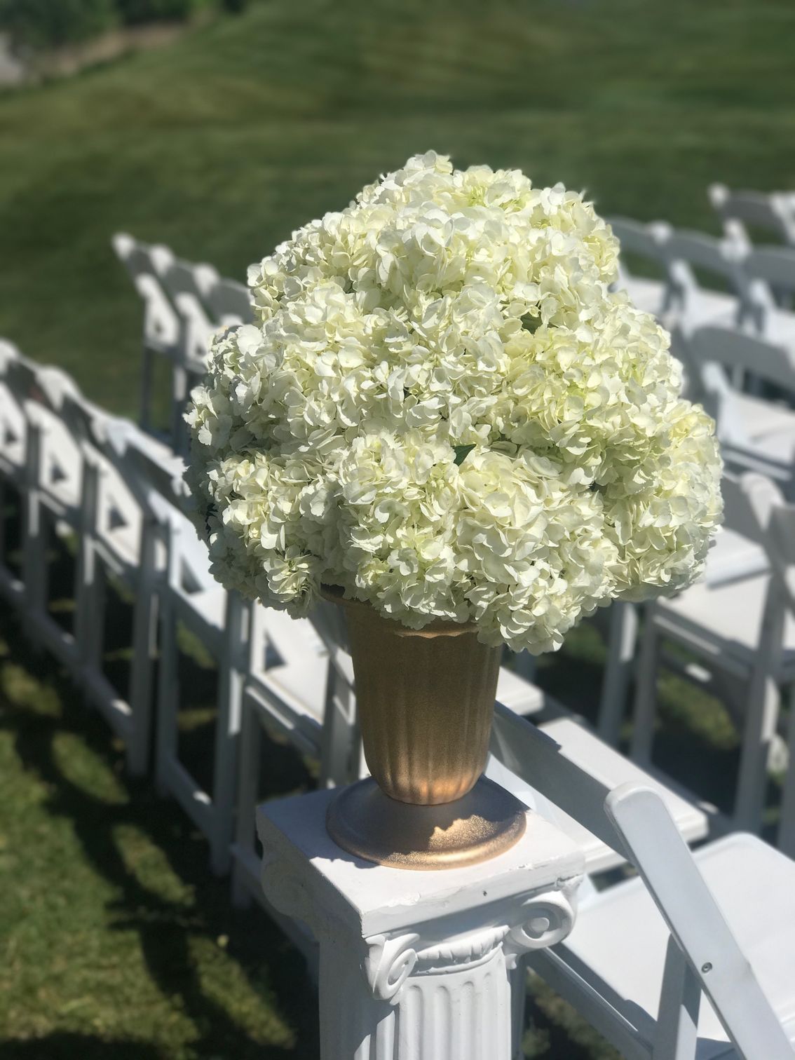 A vase filled with white flowers is sitting on a pedestal in front of a row of white chairs.