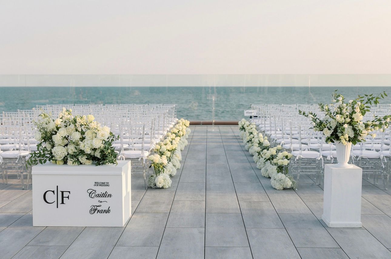 A wedding ceremony is taking place on the beach with white chairs and flowers.