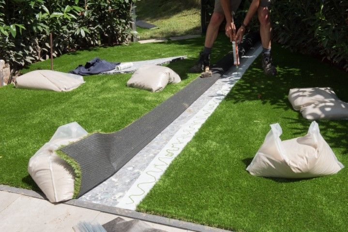 Person installing artificial turf on green lawn, holding a roll. Sandbags are placed nearby.