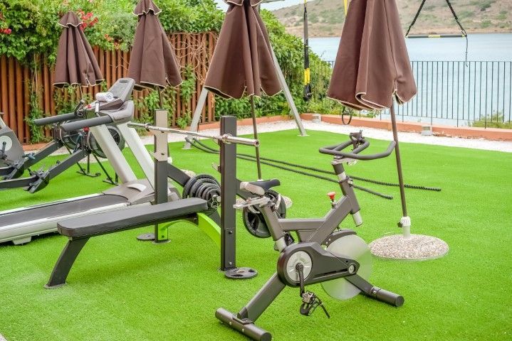 Outdoor gym with exercise equipment under brown umbrellas on green turf, with water in the background.