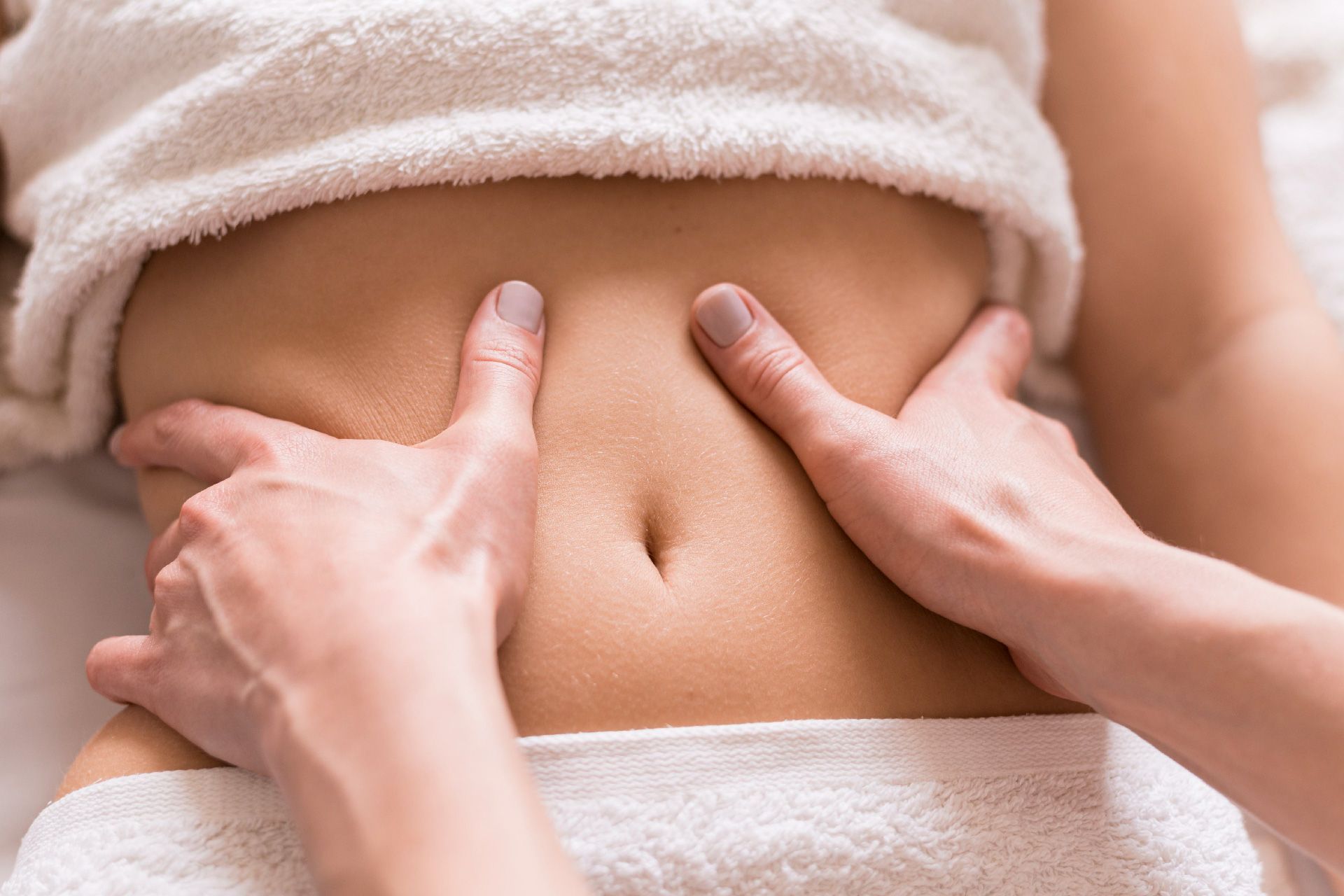 Manos masajeando el estómago desnudo de una persona, envuelto en una toalla blanca.