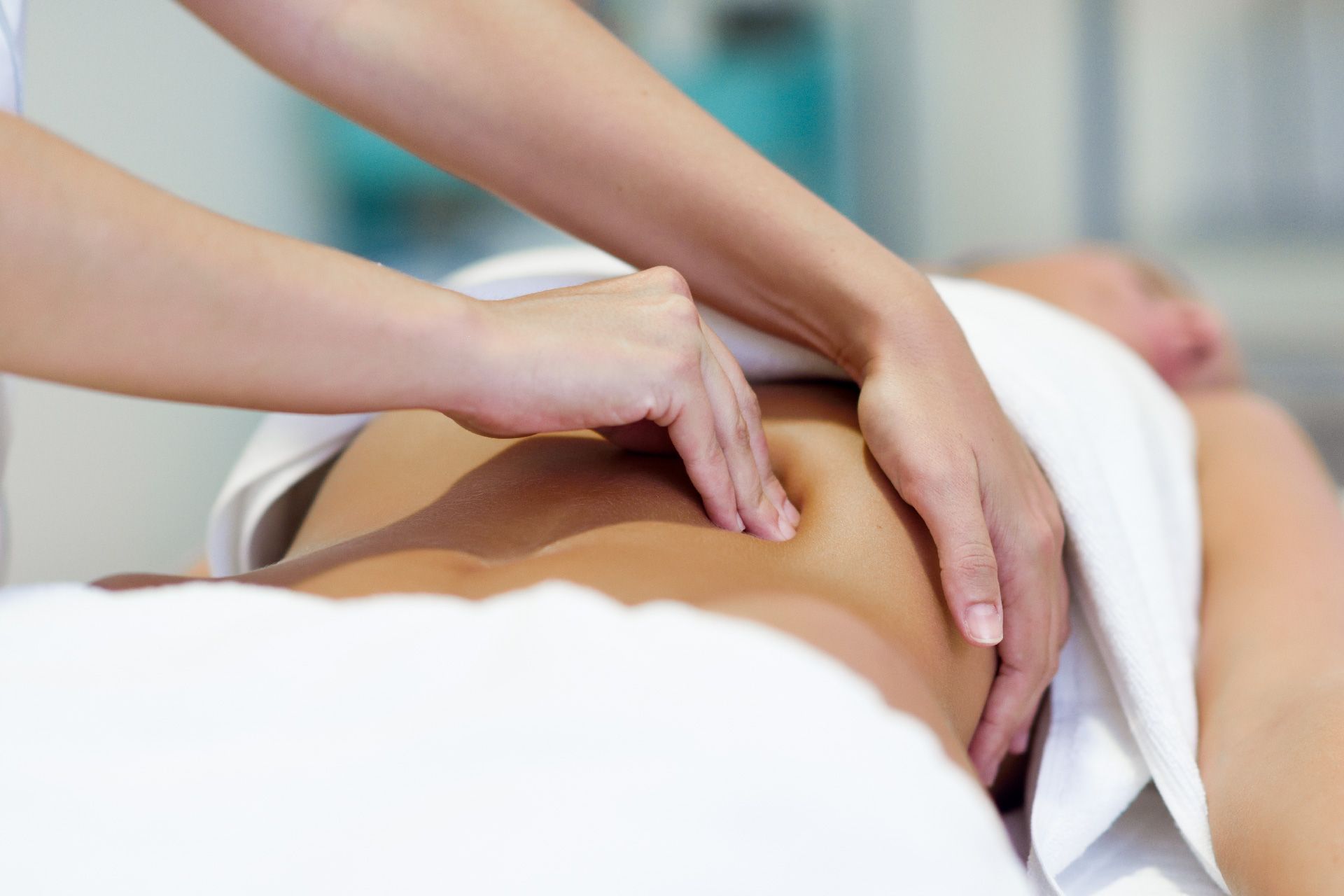 Manos masajeando la espalda baja y el abdomen de una persona, envuelta en una toalla blanca, en el interior.