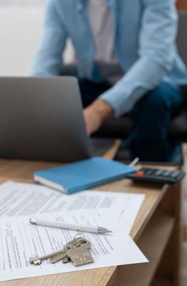 Ordenador portátil sobre el escritorio con papeles, llaves, calculadora y un cuaderno azul; persona sentada al fondo.