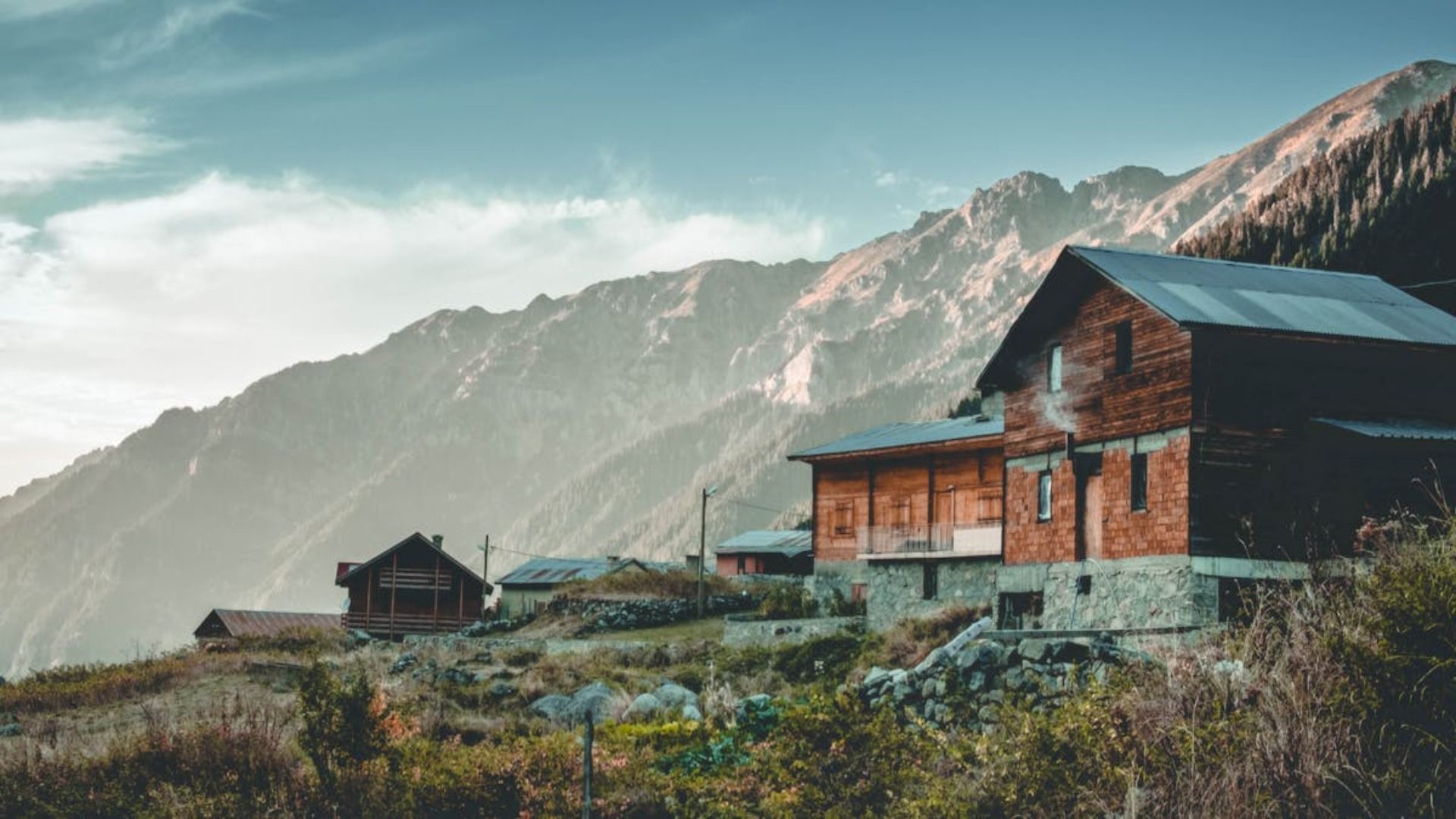 Cabañas rústicas de madera en la ladera de una montaña con picos brumosos y una suave luz matutina.