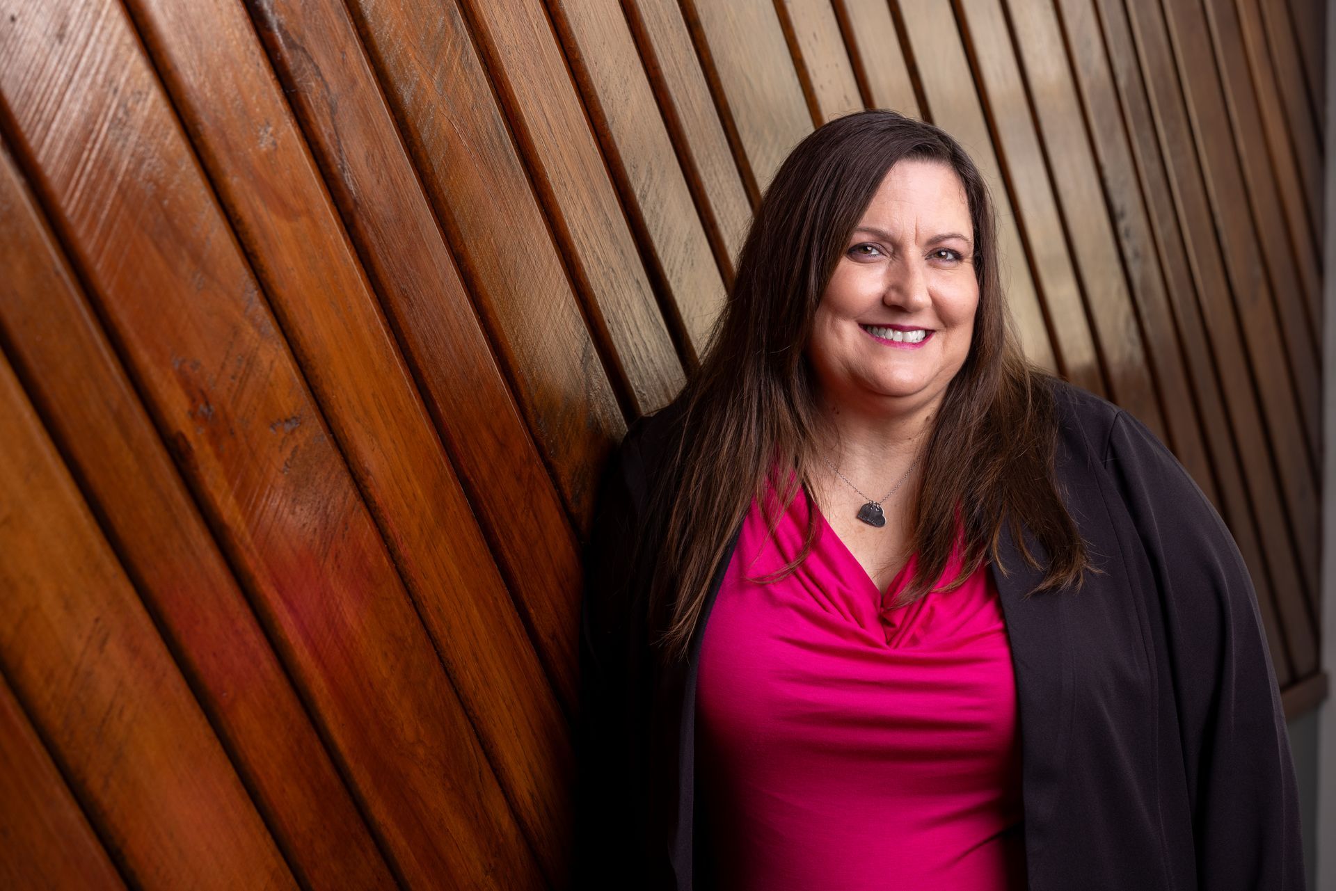 A woman in a pink shirt and black jacket is leaning against a wooden wall.