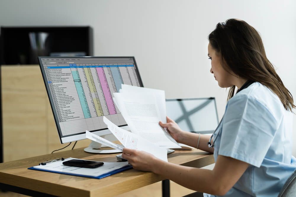 A nurse is sitting at a desk looking at papers in front of a computer.