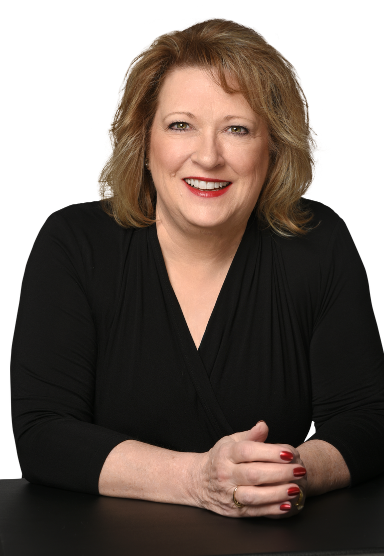 A woman is sitting at a table with her hands folded and smiling.