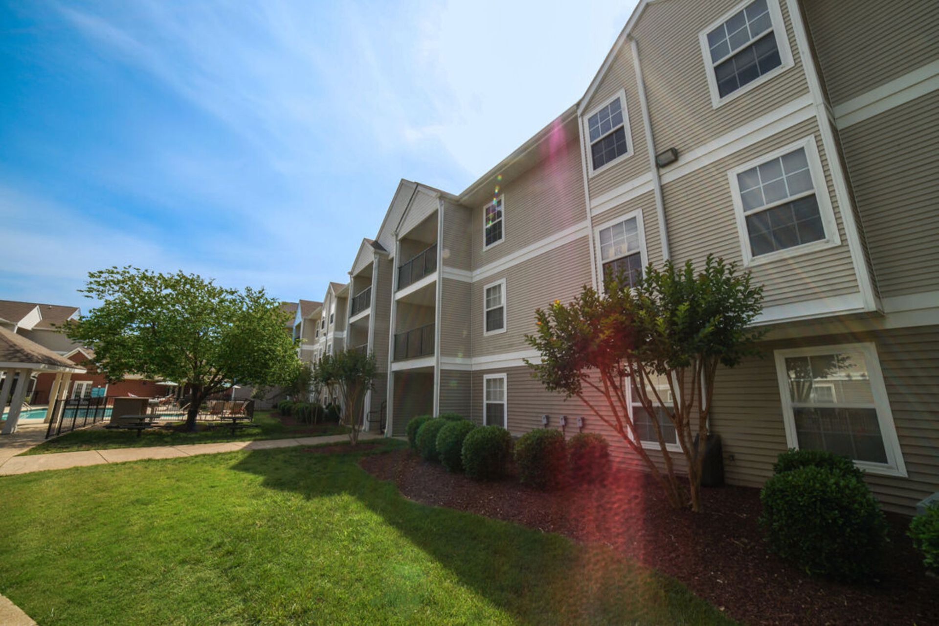 Apartment building with tan siding, balconies, and green landscaping on a sunny day.