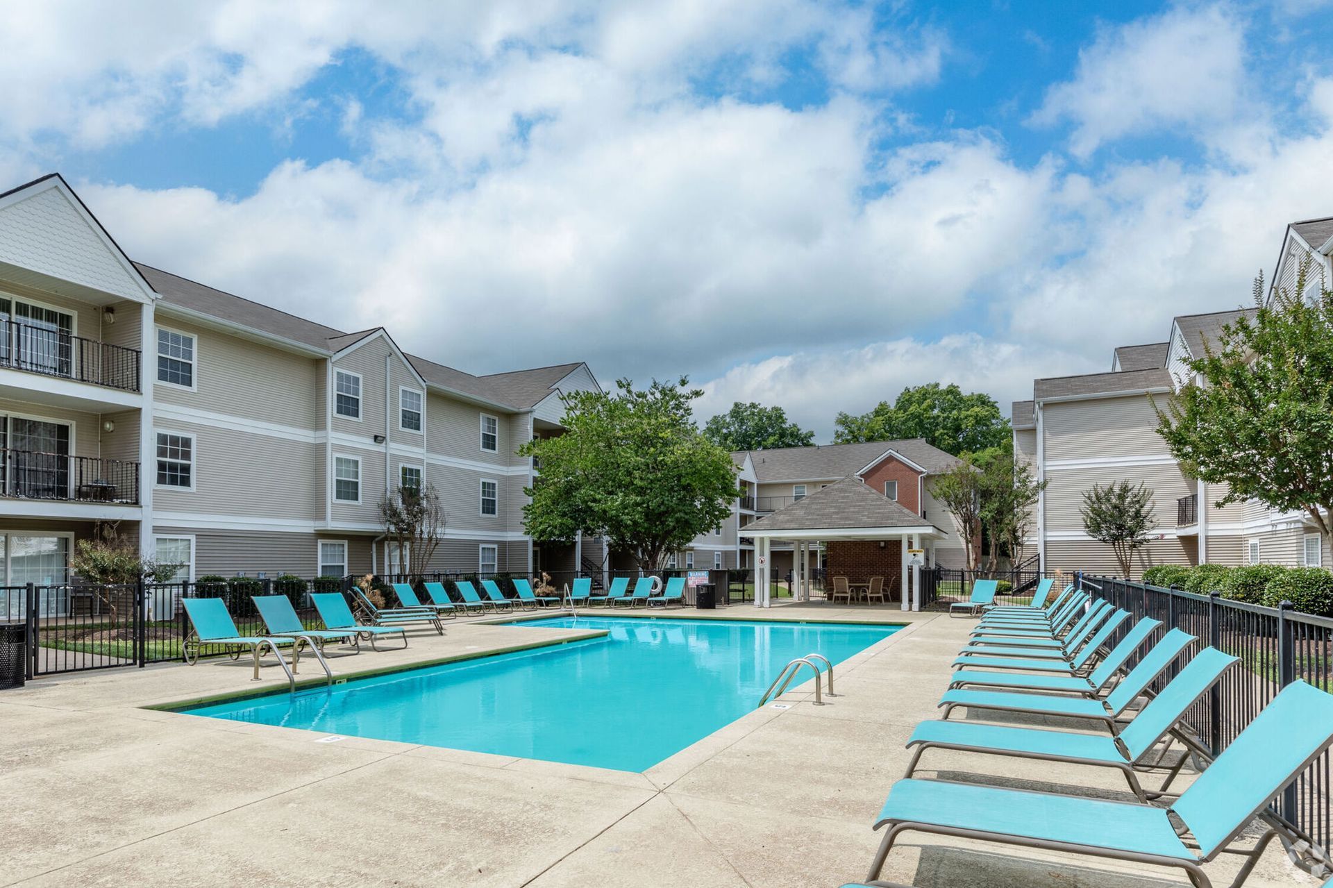 Pool with turquoise lounge chairs, apartment buildings, and a blue sky.