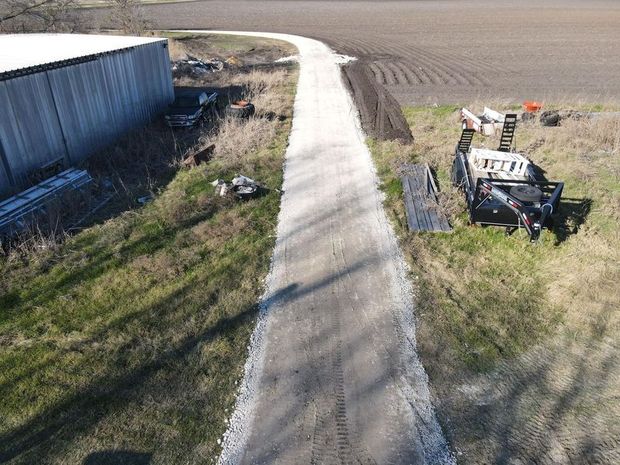 A gravel path winds through a rural field, passing a metal barn on the left and construction equipment on the right.