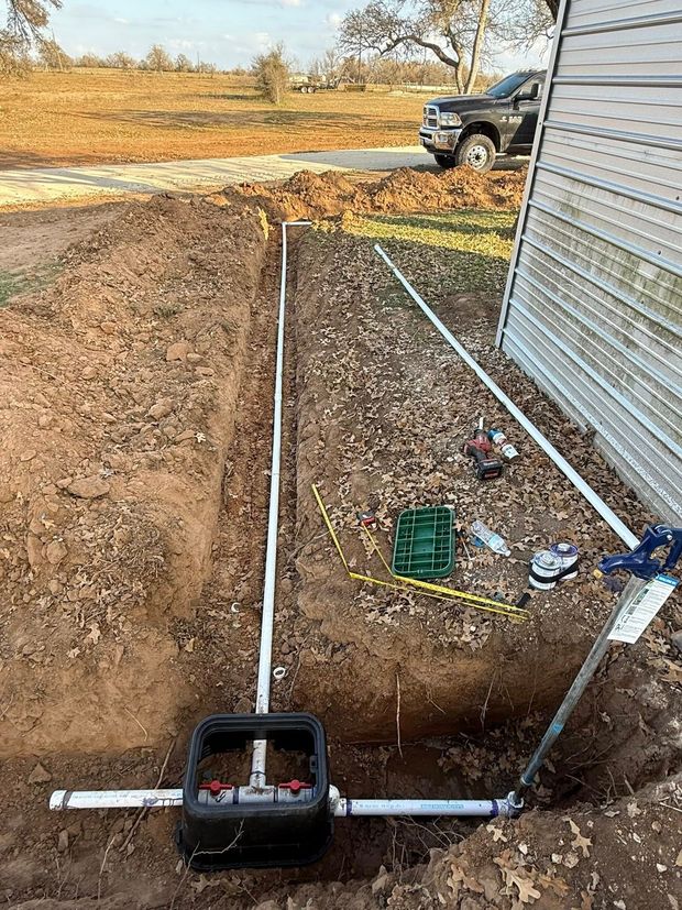 A valve box with PVC plumbing connected to irrigation lines in a dug trench next to a building.