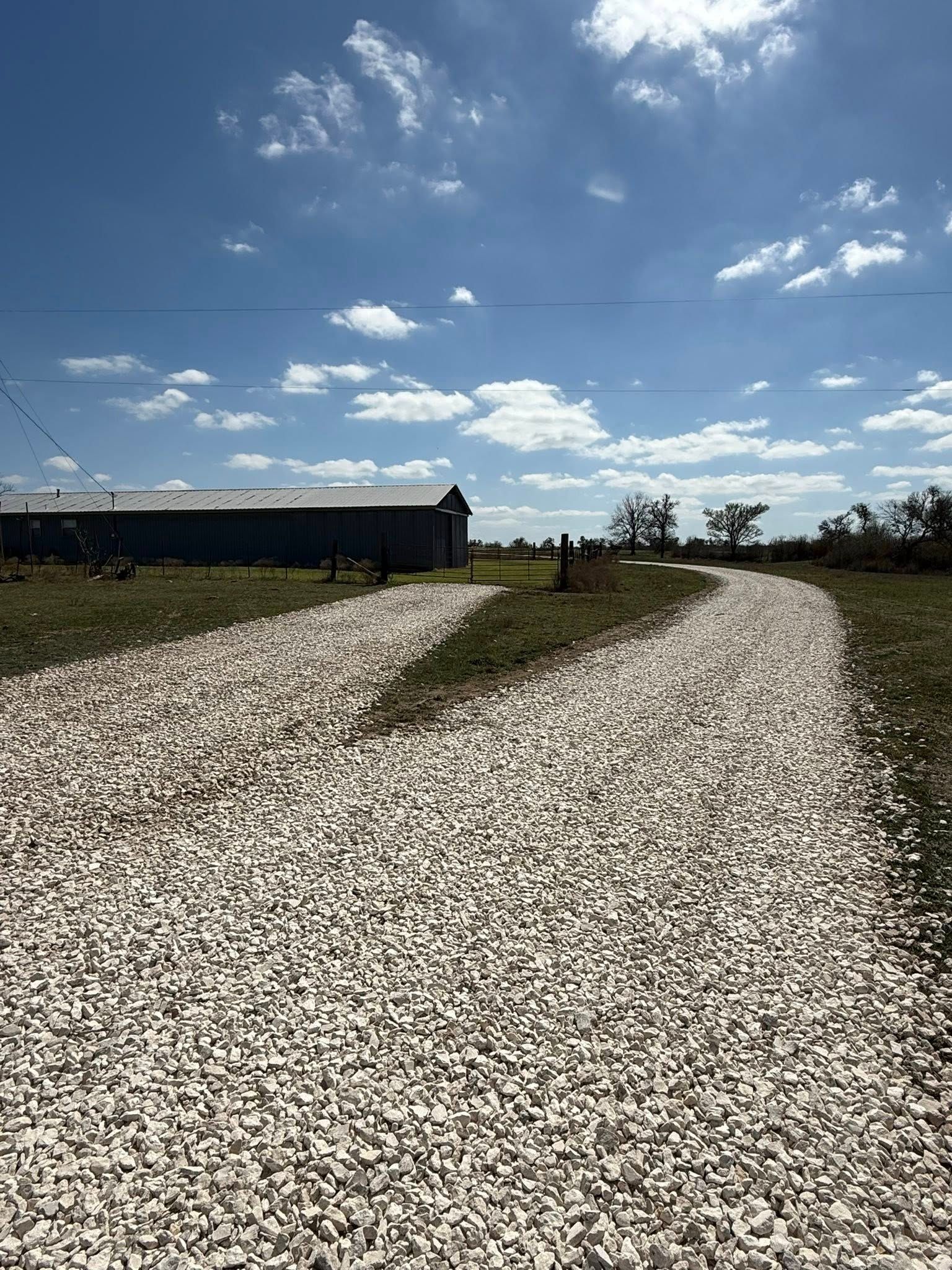 A white gravel driveway leads toward a long, dark storage building under a bright blue, partly cloudy sky.
