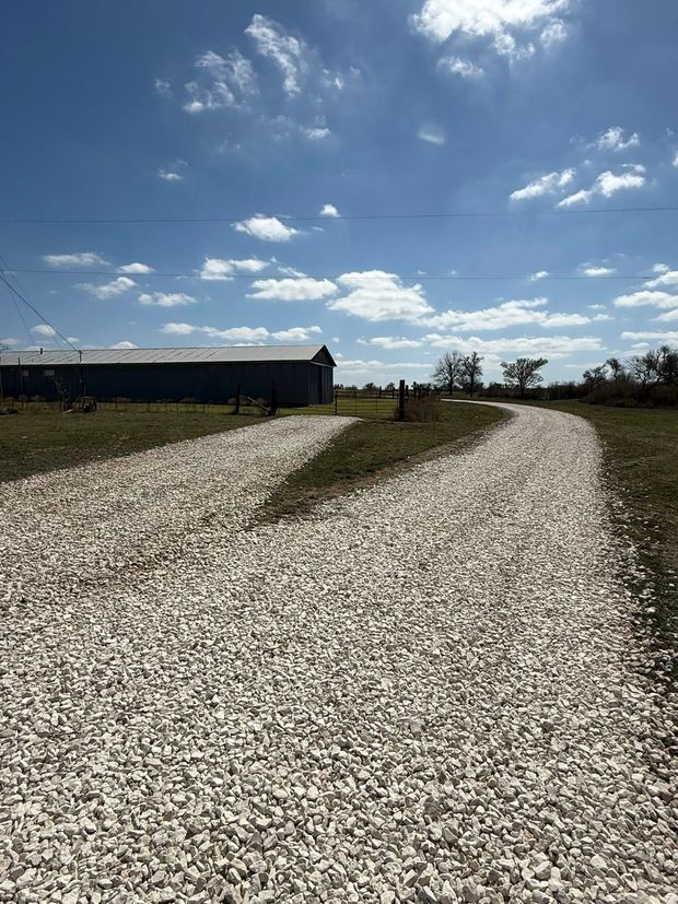 A white gravel driveway leads toward a long, dark storage building under a bright blue, partly cloudy sky.