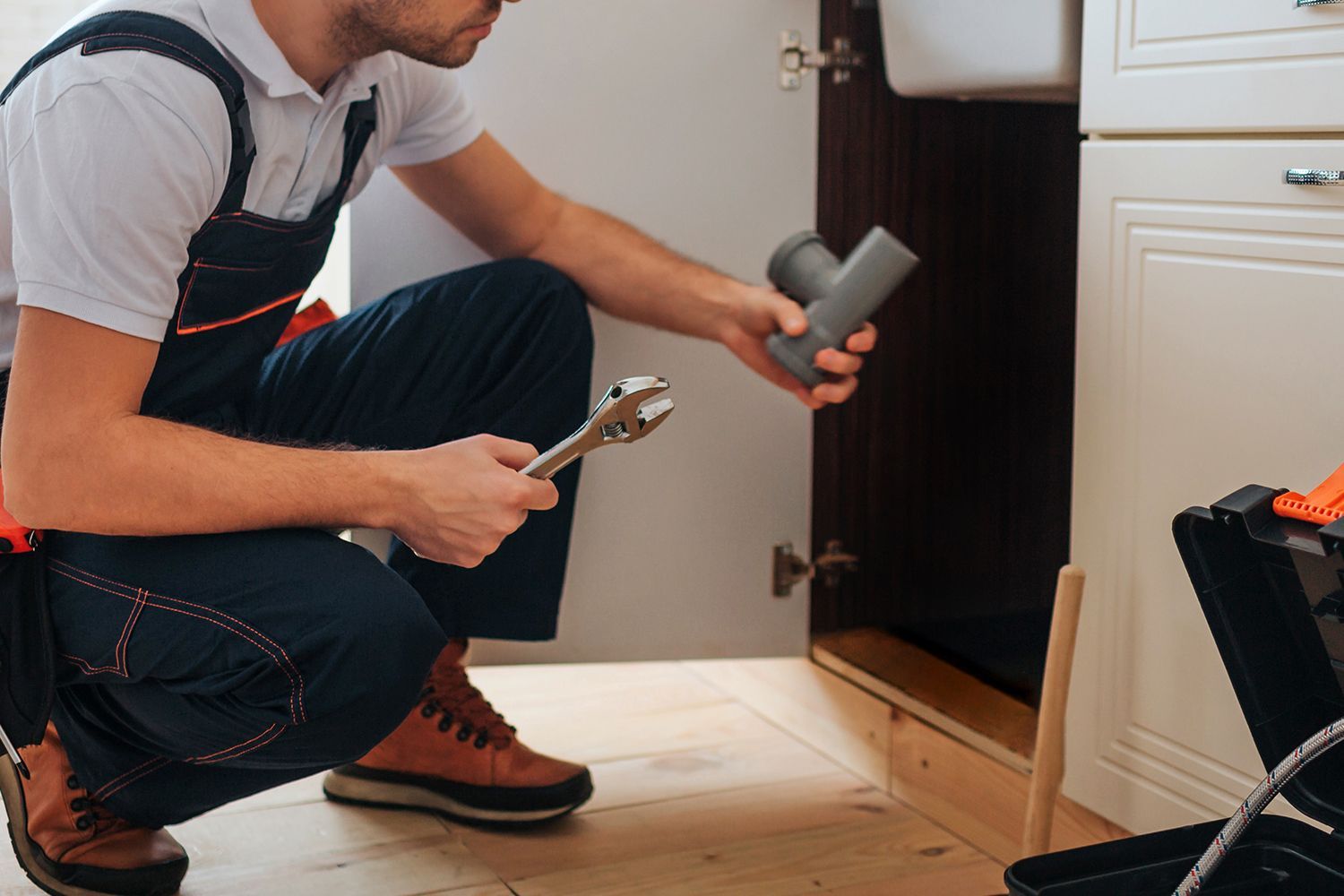 Plumber kneeling under sink holding pipe and wrench during a repair. Plumber kneeling under sink holding pipe and wrench during a repair.