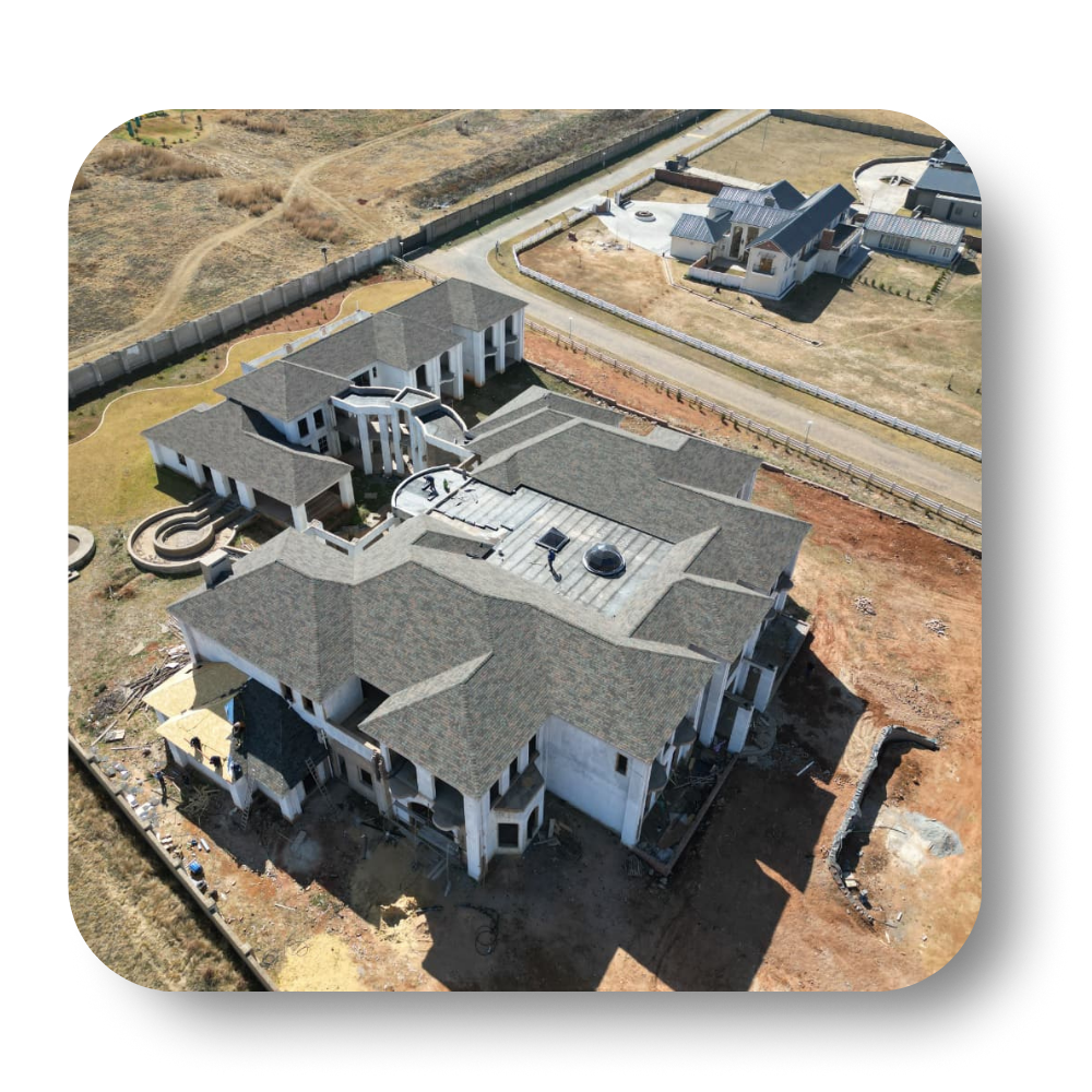 Aerial view of a large, multi-winged white house under construction, surrounded by dry land and a paved road.