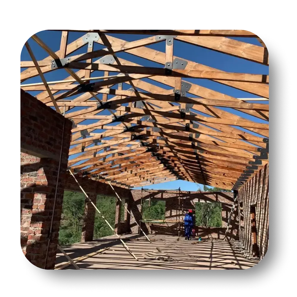 Construction of a wooden roof framework. Brick walls and blue sky visible. A worker stands on the structure.