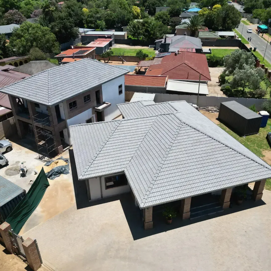 Aerial view of a building under construction with blue metal roofing and surrounding residential area with lush greenery.