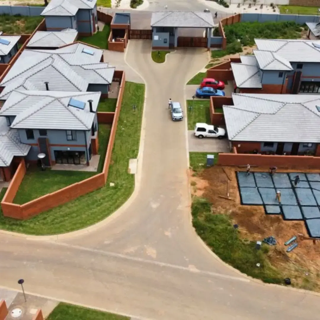 Aerial view of a residential neighborhood with gray-roofed houses and a paved road. Cars and green lawns are visible.